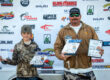 Mick and Leland holding up their first place trophies in front of sponsor banner